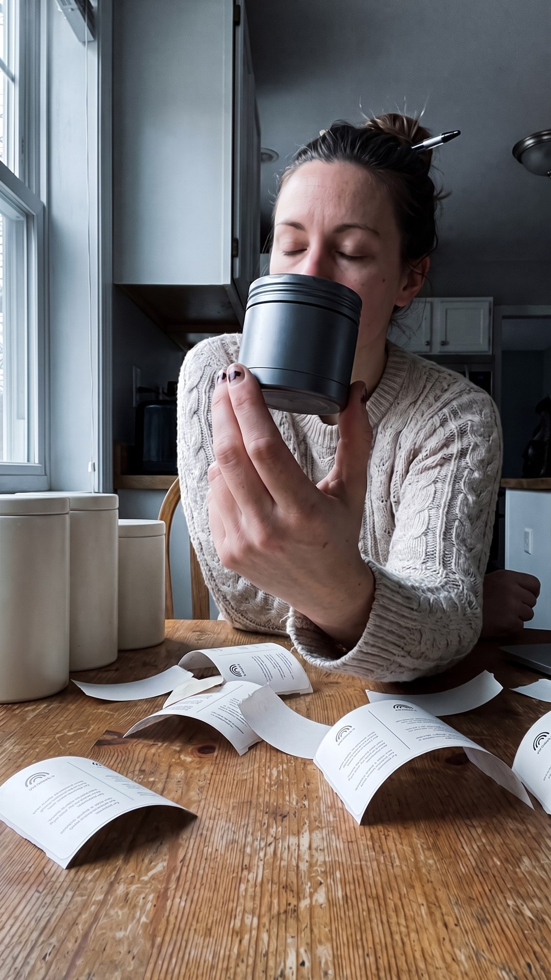 A founder closes her eyes and smells an unlabeled fragrance canister at her kitchen table, label proofs fanned out in front of her.