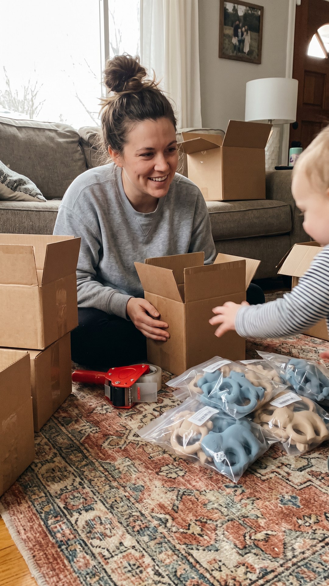 A mom sorts bagged bath-toy samples on her living room floor while her toddler reaches in to grab one.