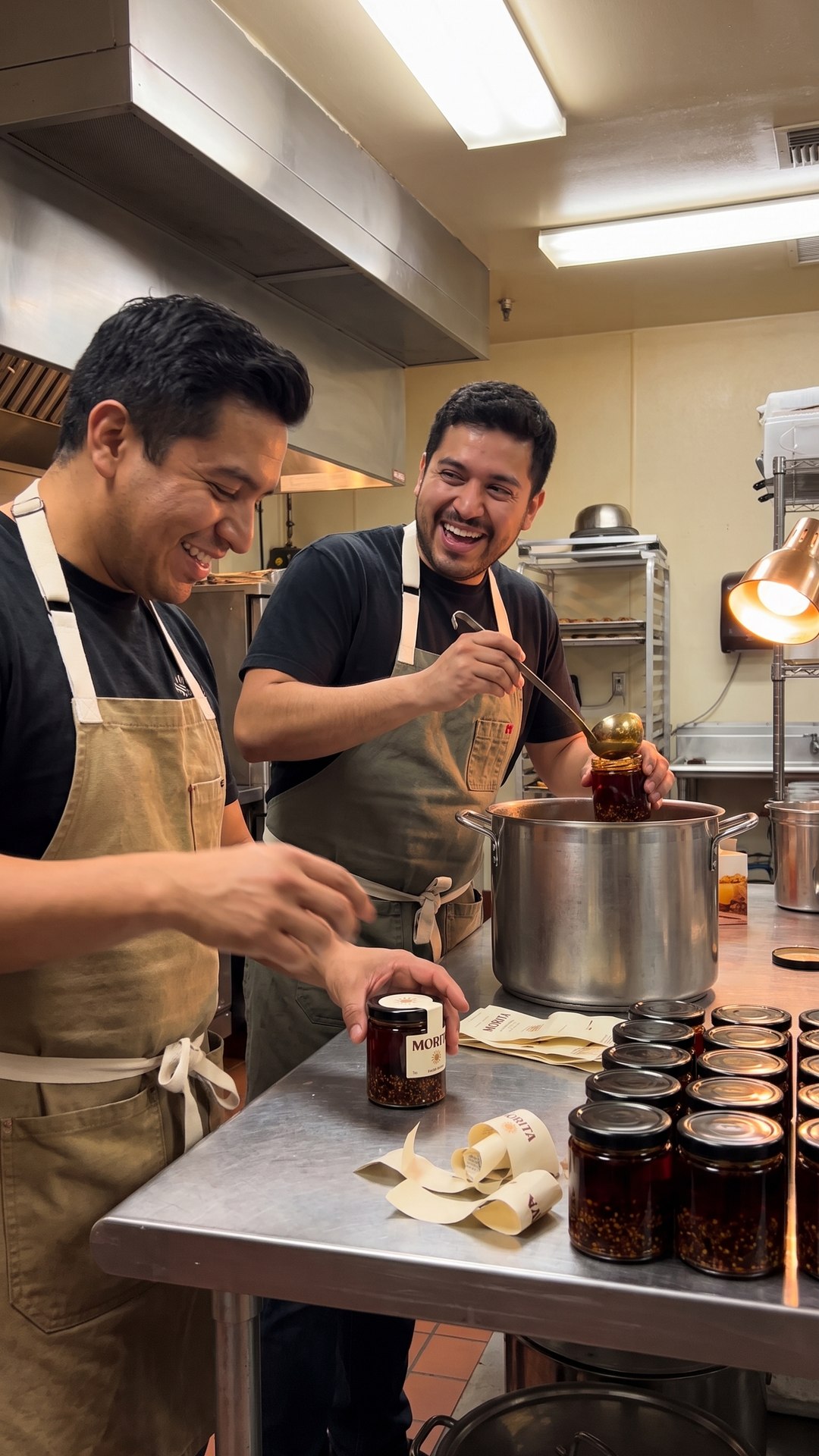 Two men laugh mid-conversation while filling and hand-labeling salsa jars in a rented commercial kitchen.