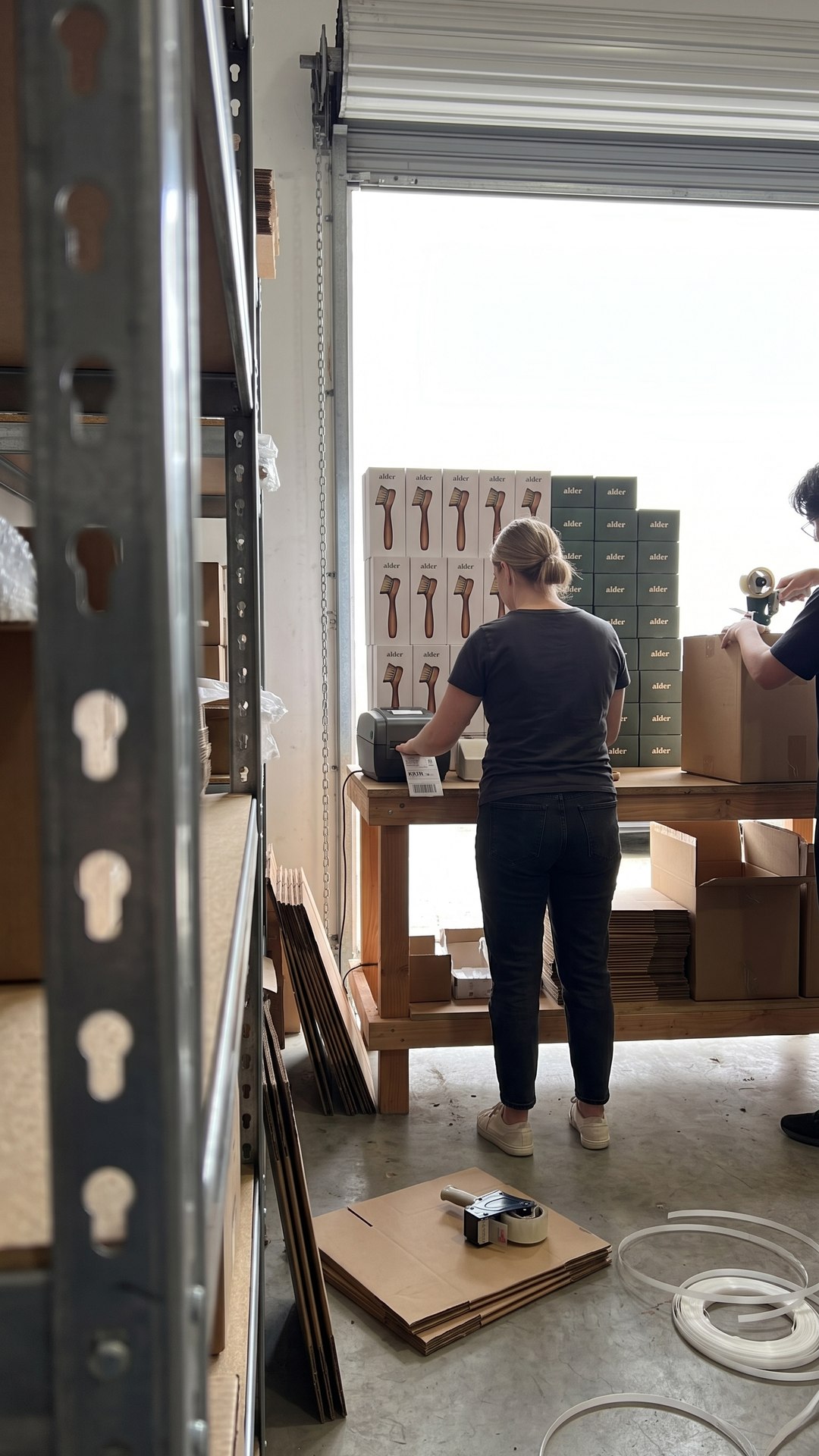 Seen from behind warehouse shelving, a founder runs a label printer at a pack-out bench stacked with branded cartons.